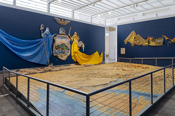 Camille Hoffman, Excelsior: Ever Upward, Ever Afloat, 2018 Queens Museum installation. Excelsior erosion control blanket; hurricane tarp; Ninja Turtles kickboard; Dollar Tree scenic landscape and golf course calendars; camouflage party napkins; tiki party decorations; Destiny inspirational poster; Lysol Clean & Fresh package; Jonah & the Whale Watchtower brochure; Planet Earth beach ball; Hudson River freight ship print-out; Thanksgiving, Tropical Paradise, Frozen, Moana, Super Bowl Sunday, Dora the Explorer, Jake the Pirate, and Finding Nemo plastic holiday tablecloths; dolphin high-pile blanket, Pioneer Supermarket, Western Beef Supermarket, Home Depot, We Care We Recycle, Thank You Thank You bodega bags, Chinatown/Ghana Must Go plastic bags; aquarium liner; stretcher bars; oil and acrylic paint on plaster, wire and wood. Courtesy of the artist. Photo by Hai Zhang.