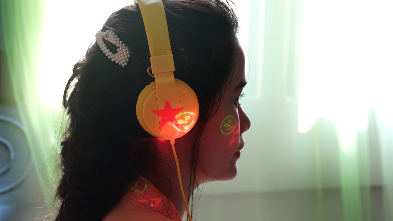 A femme person with long dark hair and yellow headphones is sitting profile to the camera. There are glowing smiley faces and red stars projected onto their face. In the background is a window, with gauzy light green curtains tinting the light coming through.