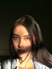A Vietnamese woman with long dark hair stands in a beam of sunlight against a nearly black background. A shadow covers her mouth.