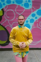 zavé martohardjono wears a long-sleeve yellow shirt with a green patterned collared shirt underneath, as well as pink pants and glasses with red frames. They are standing in front of a commercial roll-up door that has been spray painted with colorful graffiti/street art.