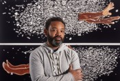 a Black man in a grey shirt with short hair and a salt-and-pepper beard stands in front of an art piece. The piece has a black background, a cluster of small white images, and a pair of Black feet and hands reach out.