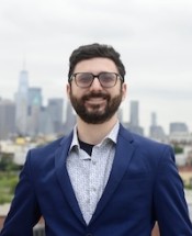 Portrait of Martin wearing a blue jacket with NYC skyline behind him