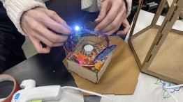 image of student's hands holding a blue, lit LED on top of a circuit assembly including a motor inside a wooden, lasercut box