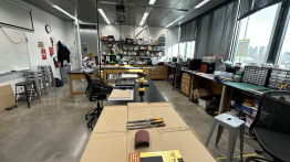 image of a makerspace/lab -there are work tables in the foreground with power tools and materials on them, and in the background shelves full of eqipment/materials and student prototypes made in the lab