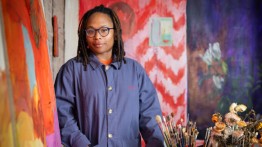 Portrait of Packer in blue jacket with painting easels and paint brushes near her