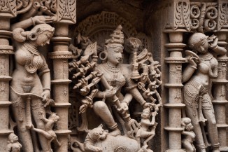 A panel detail from the Queen's Stepwell with sculptures depicting the mighty goddess Durga slaying a demon in the middle, flanked by two maidens clothing and adorning themselves with jewelry.  